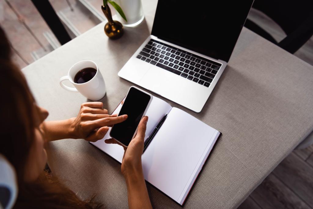 bijgesneden weergave van vrouw studeren online met smartphone en laptop in cafe met koffiekop - Foto, afbeelding
