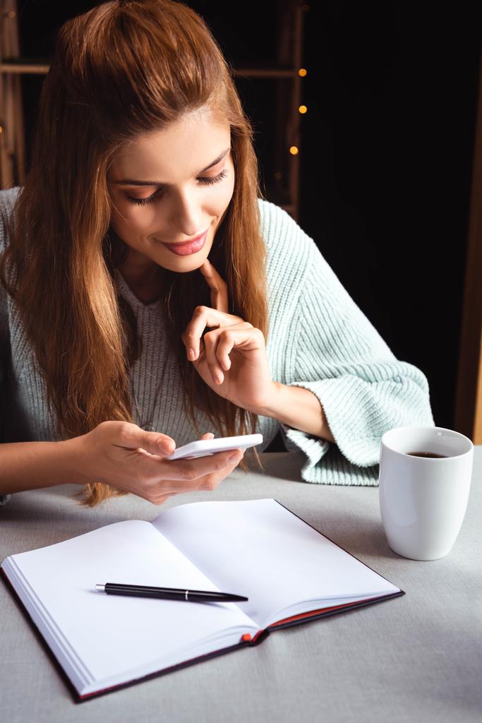 ελκυστική γυναίκα που χρησιμοποιεί smartphone στο καφέ με σημειωματάριο και καφέ  - Φωτογραφία, εικόνα