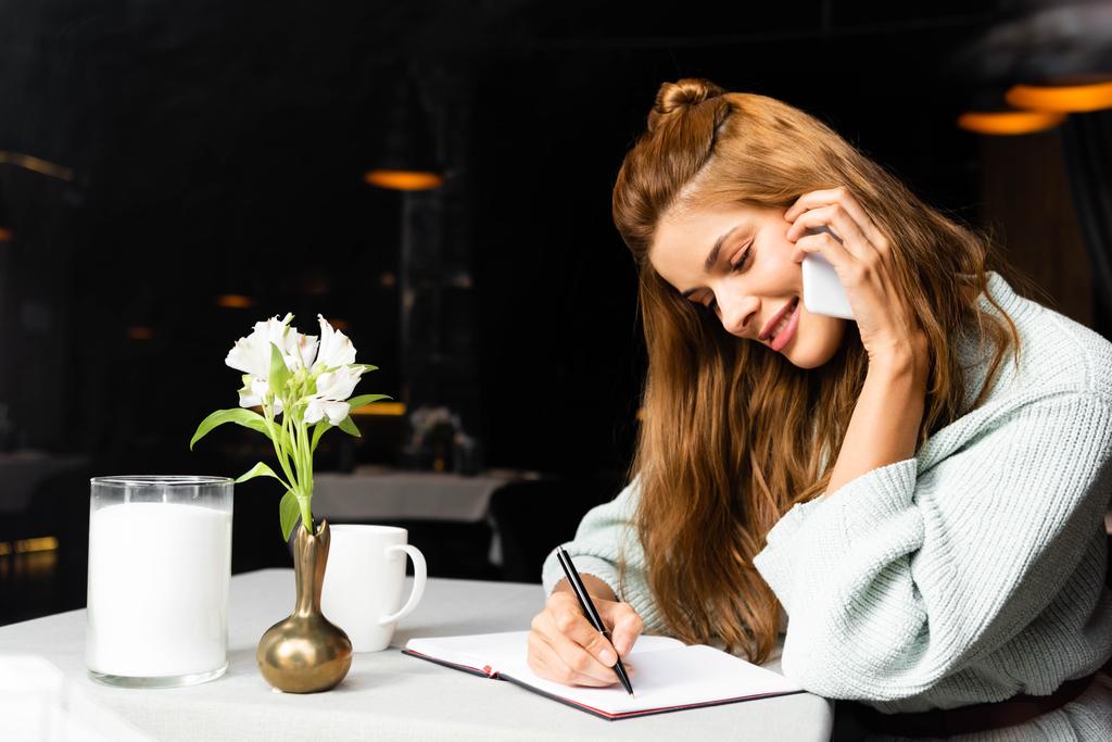 uśmiechnięta dziewczyna rozmawia na smartfonie i pisze w notatniku w kawiarni i filiżance kawy - Zdjęcie, obraz