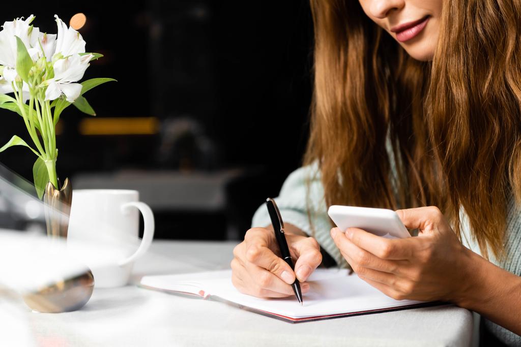 обрізаний вид жінки за допомогою смартфона і написання в блокноті в кафе з чашкою кави
 - Фото, зображення