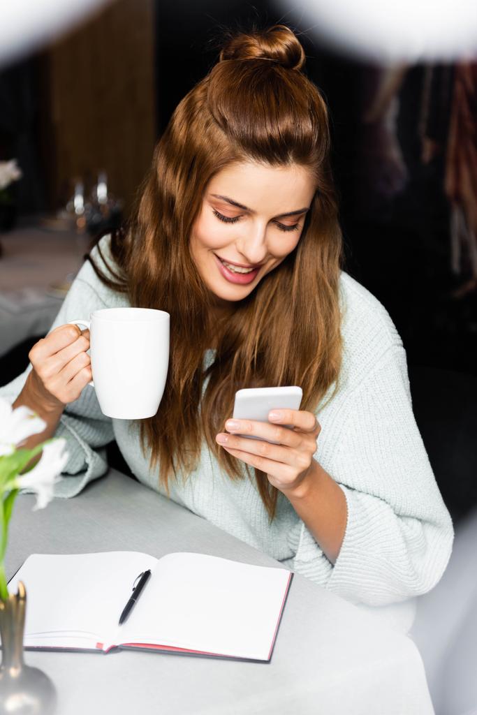 ノート付きのカフェでスマートフォンを使ってコーヒーを飲む幸せな女性  - 写真・画像