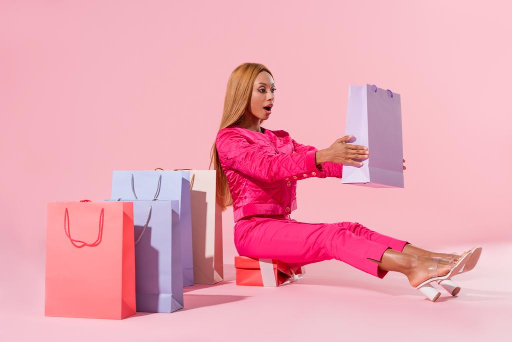 amused african american woman sitting on gift box and holding shopping bag on pink background, fashion doll concept - Photo, Image