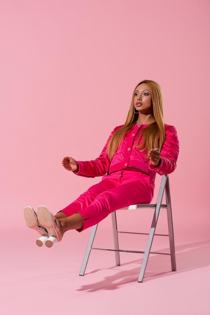 modna Afroamerykanka siedzi na krześle z rozciągniętymi nogami na różowym tle, koncepcja lalki mody - Zdjęcie, obraz