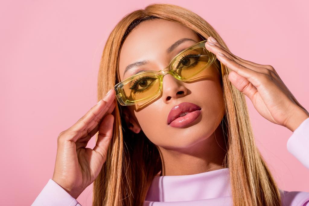 Porträt einer eleganten, blonden Afroamerikanerin, die eine Brille auf rosa, Modepuppen-Konzept berührt - Foto, Bild