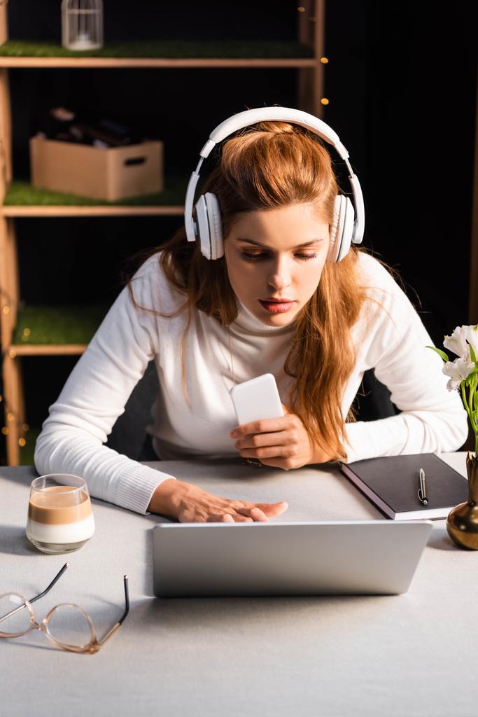 красивая сосредоточенная женщина в наушниках смотрит вебинар на ноутбуке в кафе со смартфоном
 - Фото, изображение
