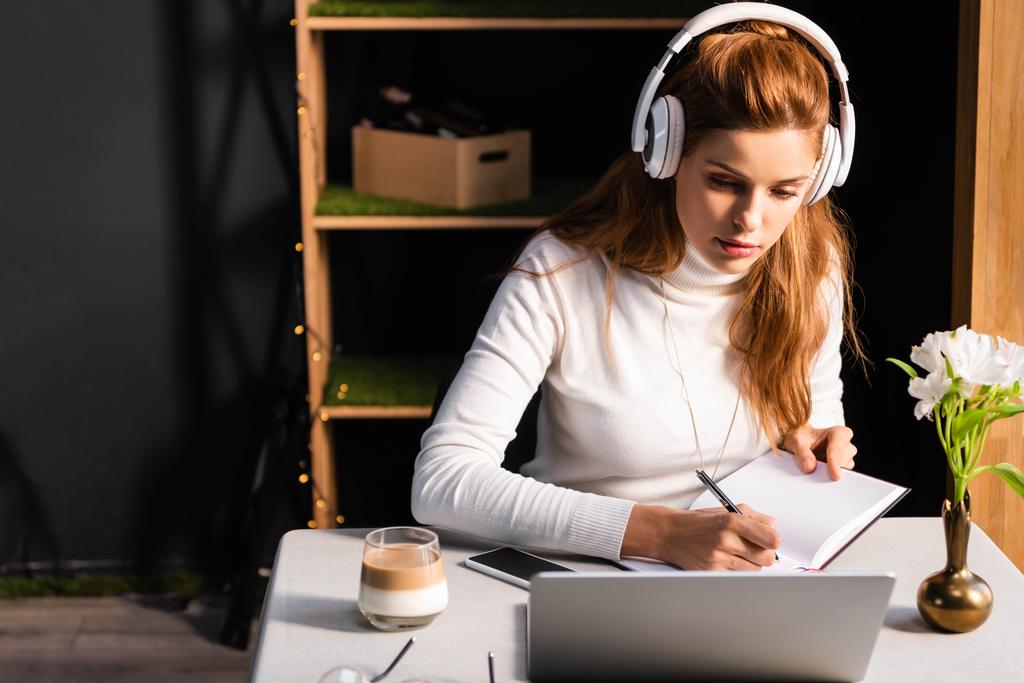 belle rousse femme dans les écouteurs écriture dans le bloc-notes tout en regardant webinaire sur ordinateur portable dans le café
 - Photo, image
