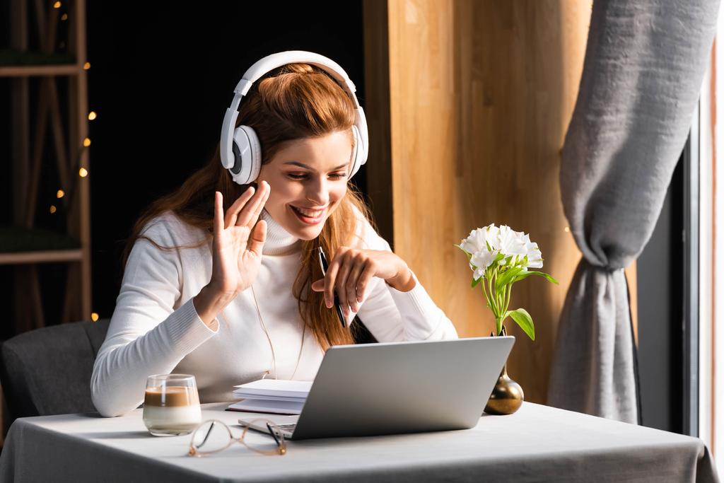 красива щаслива жінка в навушниках махає і веде відео чат на ноутбуці в кафе
 - Фото, зображення