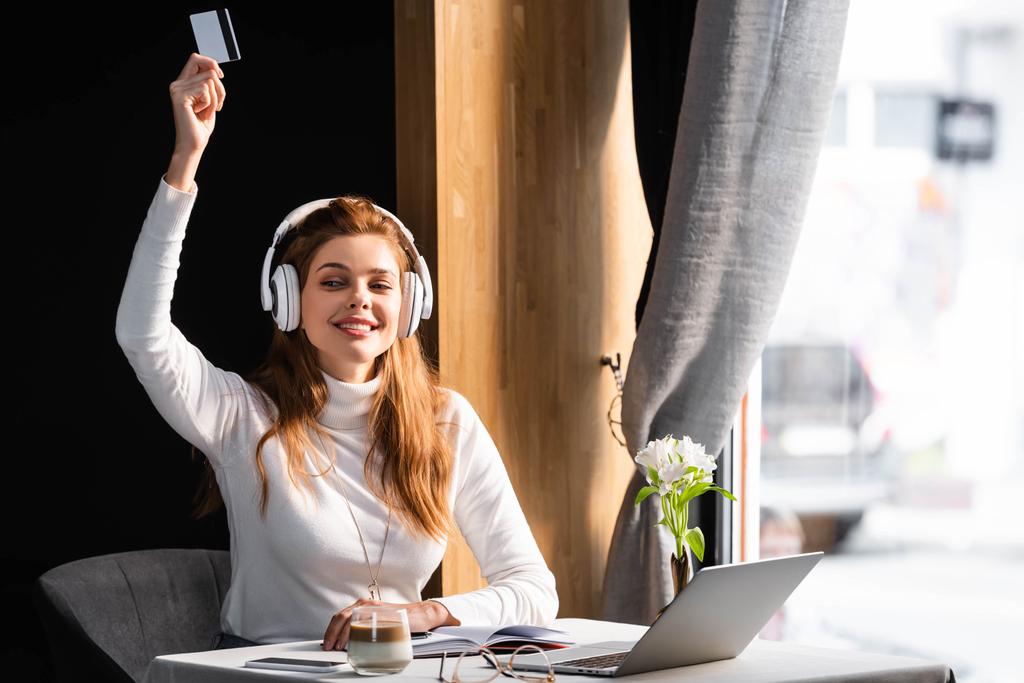 krásná zrzka žena ve sluchátkách placení kreditní kartou v kavárně s notebookem, notebookem a kávou - Fotografie, Obrázek