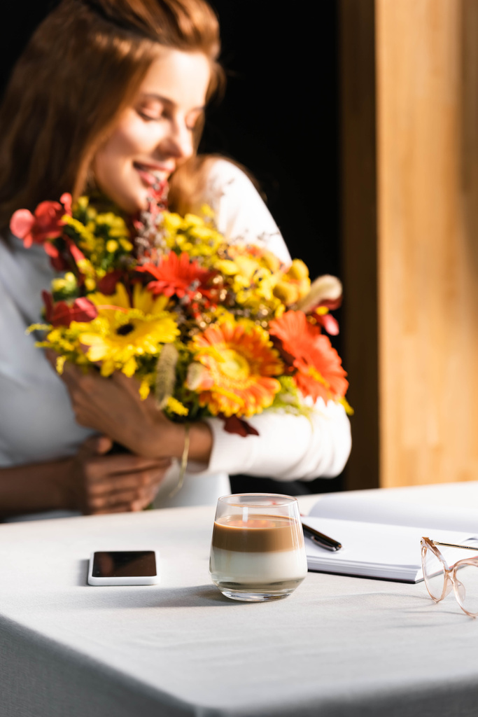 enfoque selectivo de la mujer feliz con ramo de flores de otoño en la cafetería con taza de café, teléfono inteligente y portátil
 - Foto, Imagen