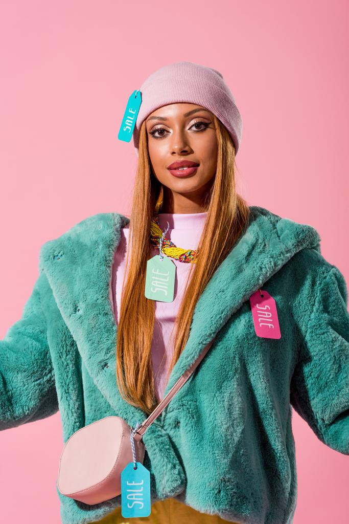 ピンクで隔離されたカメラを見て服の販売ラベルを持つスタイリッシュなアフリカ系アメリカ人女性,ファッション人形の概念 - 写真・画像