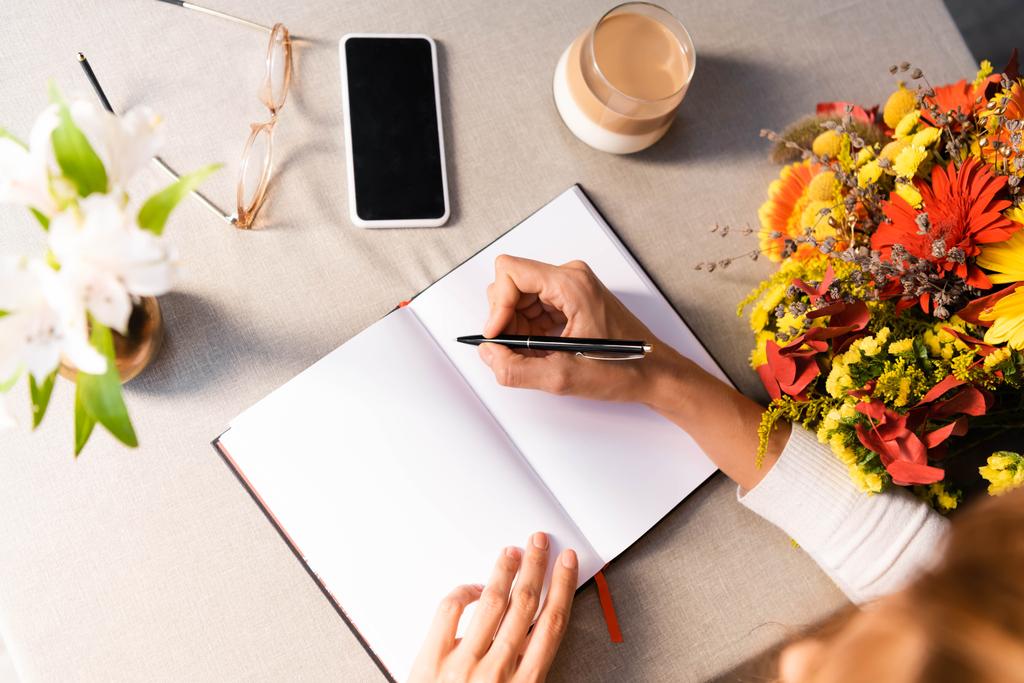 обрізаний вид жінки, що пише в блокноті в кафе з кавою, смартфоном і квітами
  - Фото, зображення
