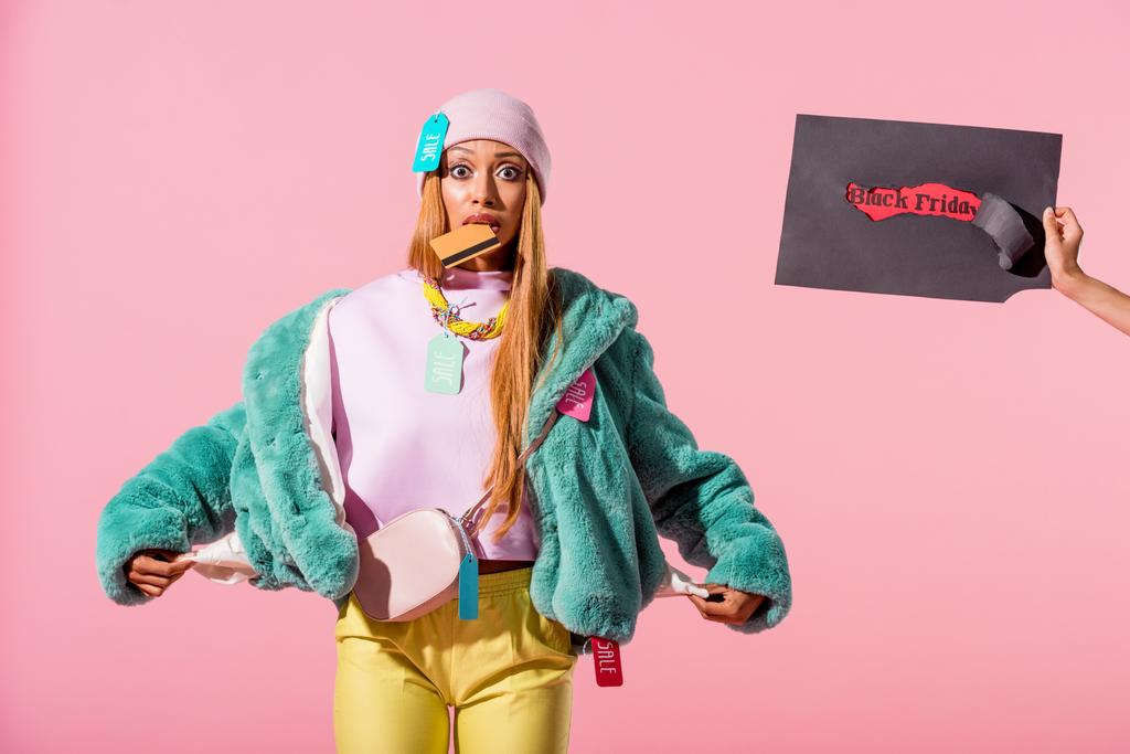 cropped view of woman holding torn paper with black friday inscription near african american woman showing empty pockets isolated on pink, fashion doll concept - Photo, Image