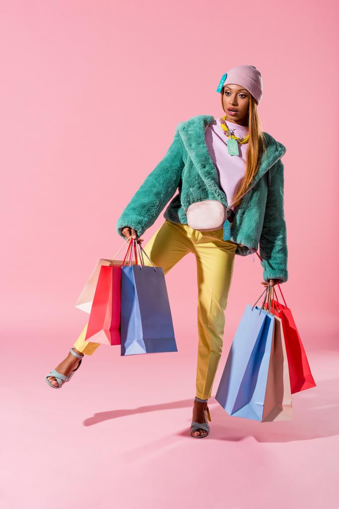 ελκυστική, κομψή αφρικάνικη αμερικανική γυναίκα κρατώντας τσάντες ψώνια, ενώ στέκεται στο ένα πόδι σε ροζ φόντο, μόδα κούκλα έννοια - Φωτογραφία, εικόνα