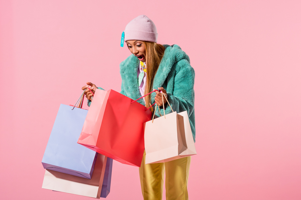 amüsiert, trendige afrikanisch-amerikanische Frau sucht in Einkaufstasche auf rosa Hintergrund, Modepuppenkonzept - Foto, Bild