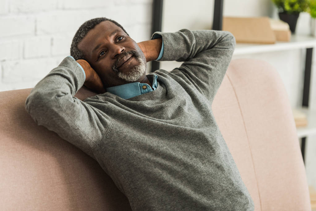 verträumter, positiver afrikanisch-amerikanischer Mann schaut weg, während er auf dem Sofa sitzt - Foto, Bild