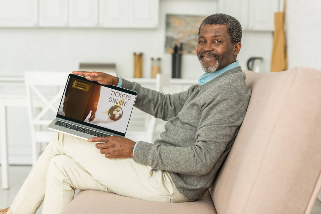 smiling african american man sitting on sofa and showing laptop with tickets online app on screen - Photo, Image