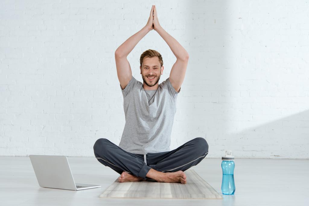 homem sorrindo olhando para a câmera enquanto pratica meia postura de lótus com mãos de oração levantadas perto de laptop e garrafa de esportes
 - Foto, Imagem