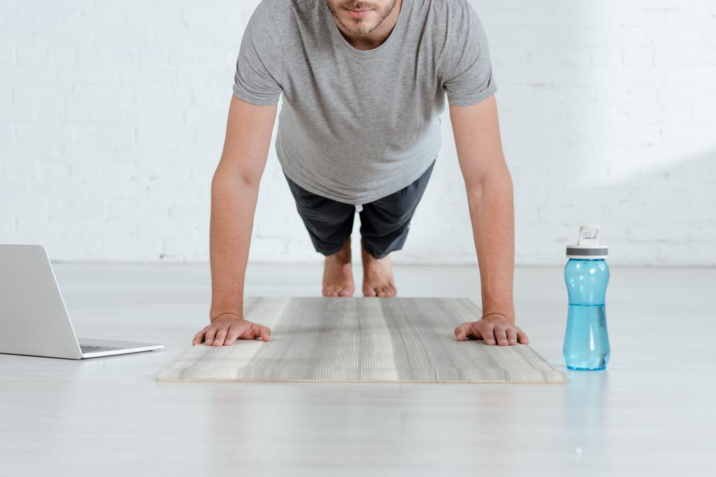 gedeeltelijke weergave van de mens oefenen onderarm plank pose in de buurt van laptop en sportfles - Foto, afbeelding