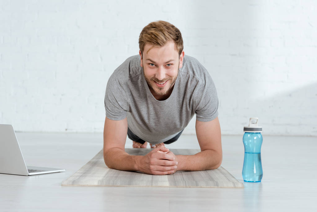glimlachende man kijken naar de camera tijdens het oefenen van onderarm plank pose in de buurt van laptop en sportfles - Foto, afbeelding