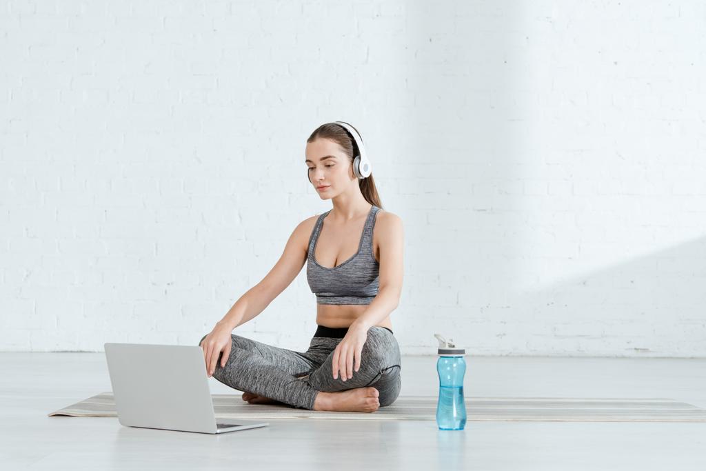 junge Frau mit Kopfhörern sitzt in lockerer Pose neben Laptop und Sportflasche - Foto, Bild