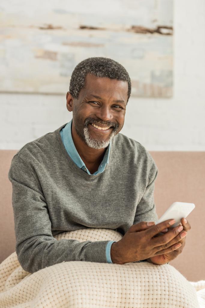 vrolijk Afrikaans amerikaanse man zitten met deken op knieën, met smartphone en glimlachen op de camera  - Foto, afbeelding
