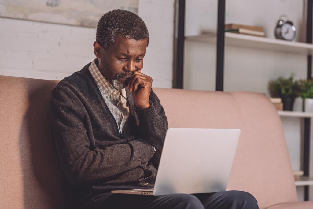 hombre afroamericano molesto mirando el ordenador portátil mientras está sentado en el sofá
 - Foto, imagen