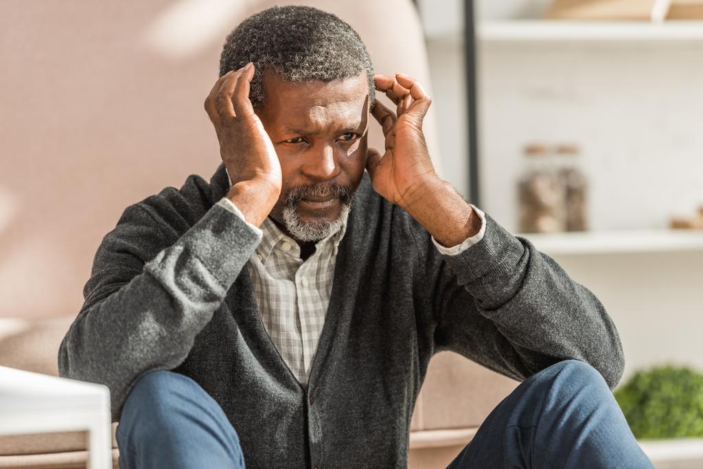 Aufgebrachter afrikanisch-amerikanischer Mann sitzt auf dem Boden und hält Hände in Kopfnähe, während er an Migräne leidet - Foto, Bild