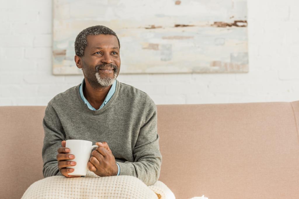 ανώτερος, θετικός Αφροαμερικάνος κάθεται με την κουβέρτα στα γόνατα και κρατώντας ένα φλιτζάνι ζεστό ποτό - Φωτογραφία, εικόνα