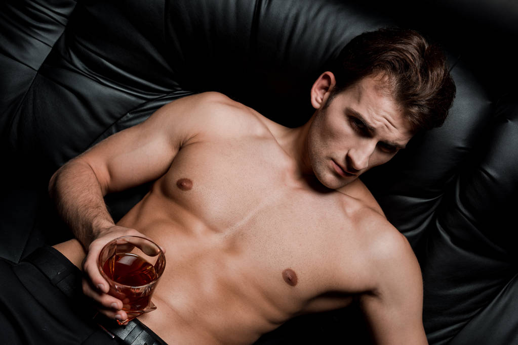 hombre guapo sentado en el sofá con un vaso de whisky en la habitación oscura
 - Foto, Imagen