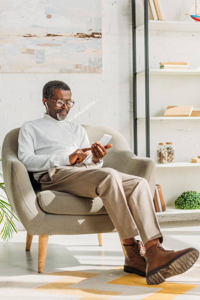 κομψό αφροαμερικανός άνδρας κάθεται στην πολυθρόνα, ακούγοντας μουσική σε τηλέφωνα και χρησιμοποιώντας ψηφιακή ταμπλέτα - Φωτογραφία, εικόνα