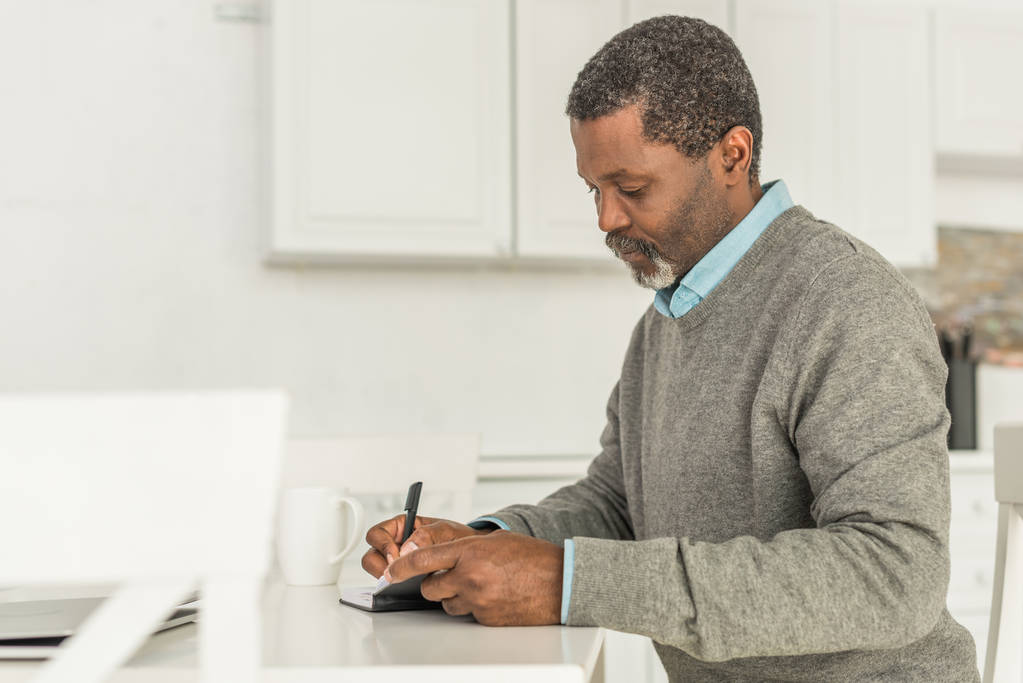 Серйозний афроамериканець сидить за столом і пише в зошиті - Фото, зображення