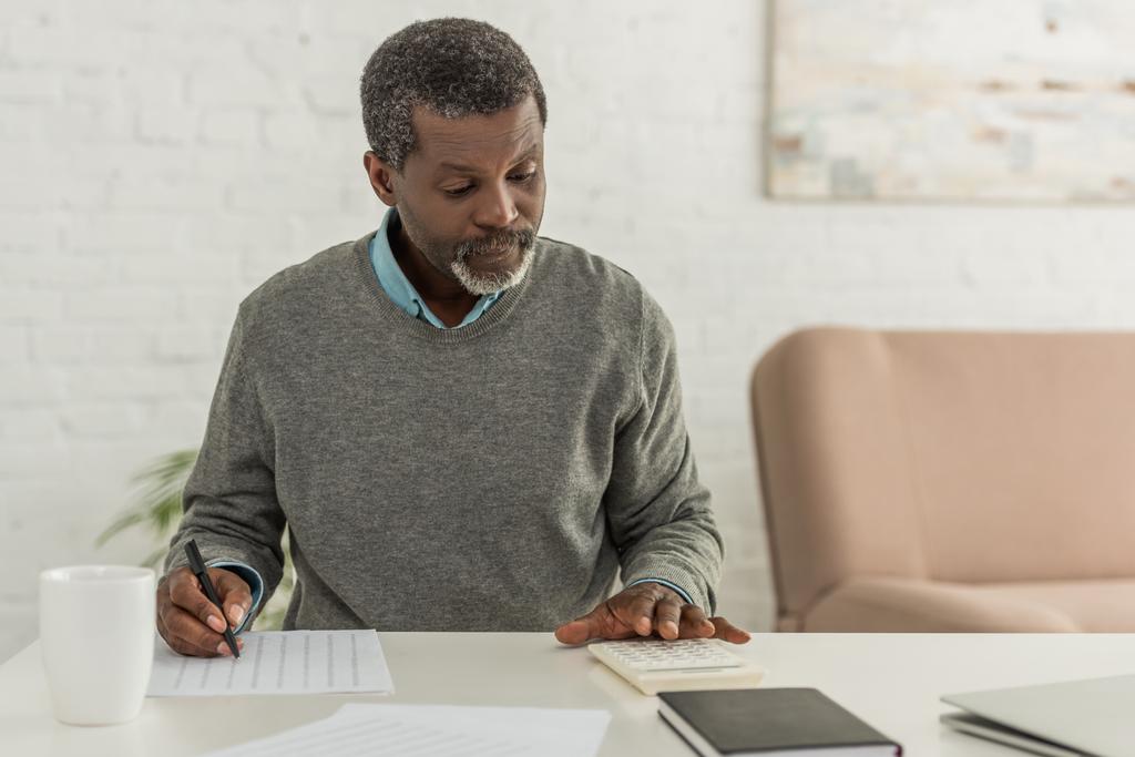 hombre afroamericano concentrado calculando gastos mientras escribe en factura de servicios públicos
 - Foto, imagen