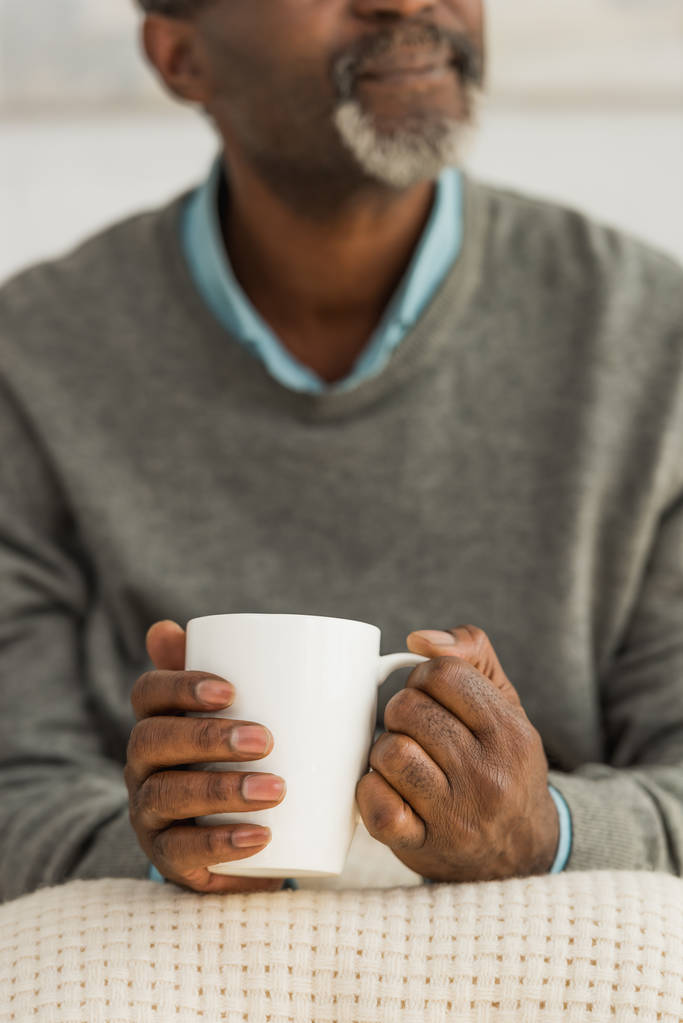 vista recortada del hombre afroamericano senior sentado con manta en las rodillas y sosteniendo la taza con bebida caliente
 - Foto, imagen