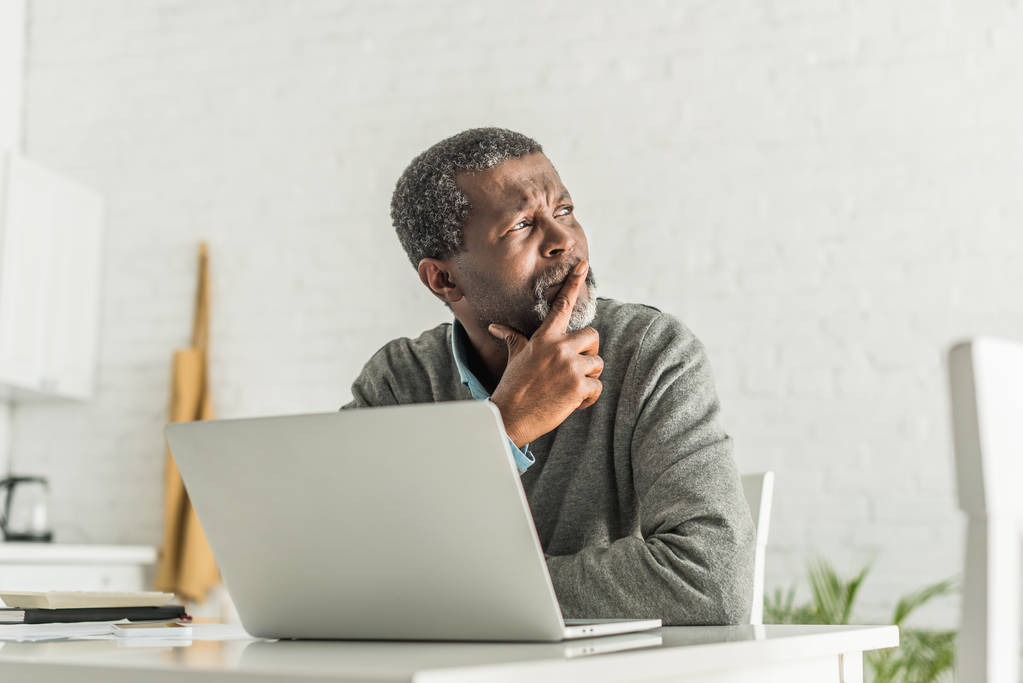 Düşünceli Afrikalı Amerikalı adam dizüstü bilgisayarın yanında otururken gözlerini kaçırıyor. - Fotoğraf, Görsel
