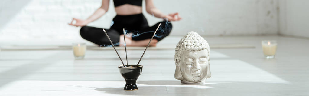 cropped view of woman sitting in half lotus pose near buddha head sculpture, aromatic sticks and candles, panoramic shot - Photo, Image