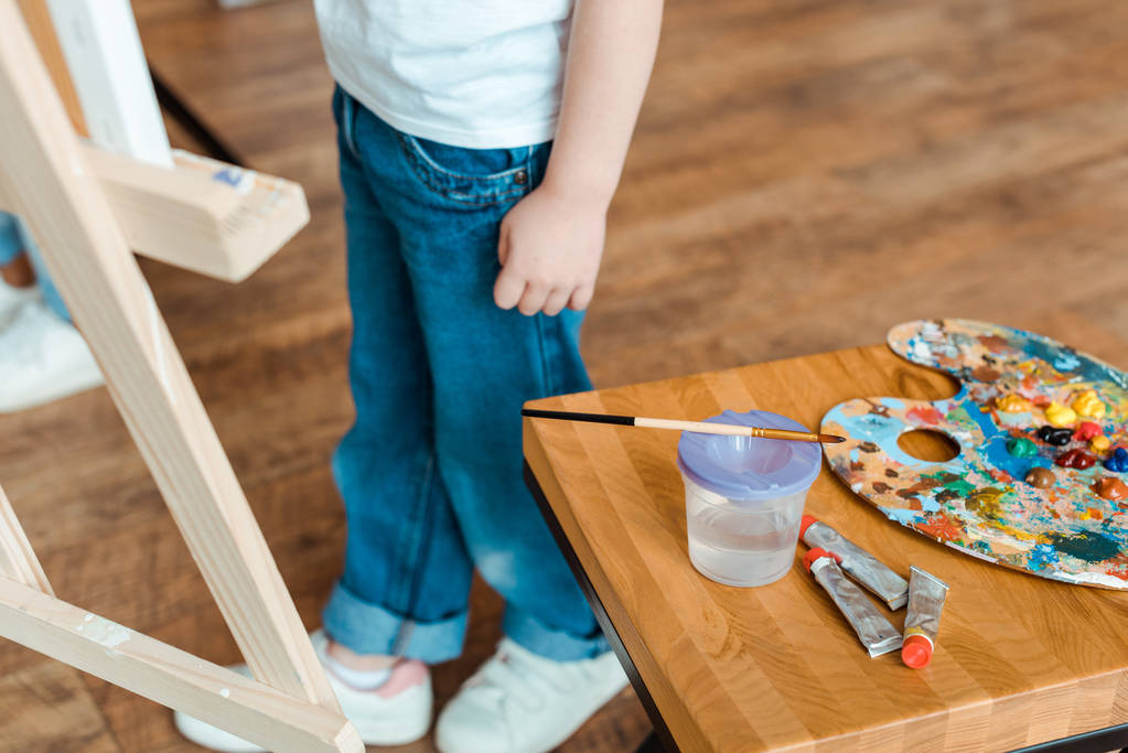 Ausgeschnittene Ansicht eines Kindes, das neben einem Holztisch mit Gouachefarben steht  - Foto, Bild