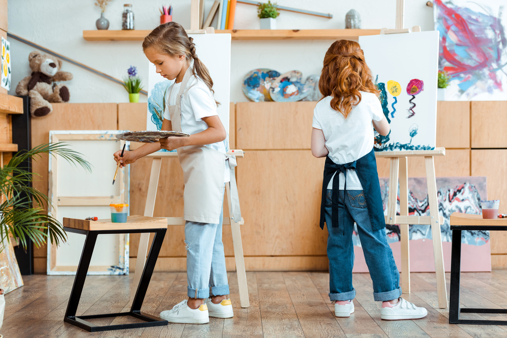 vista posteriore di rossa bambino tenendo tavolozza mentre la pittura su tela vicino bambino nella scuola d'arte
  - Foto, immagini