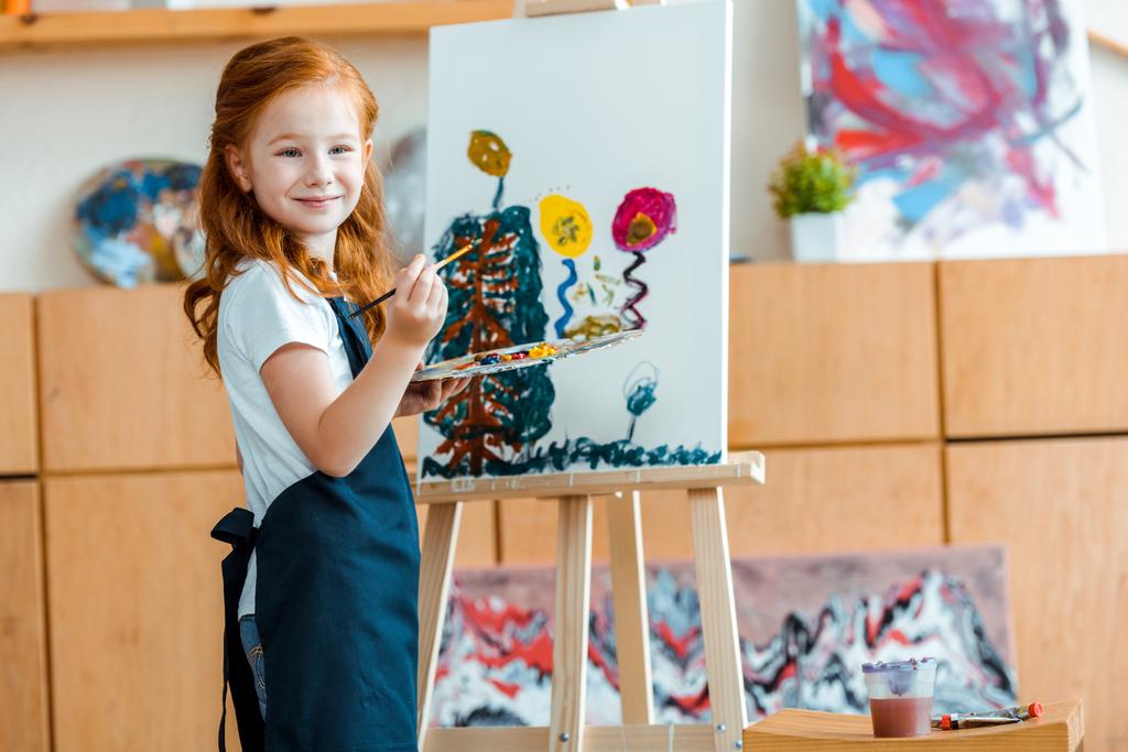 χαρούμενο κοκκινομάλλικο παιδί ζωγραφίζει σε καμβά στη σχολή καλών τεχνών  - Φωτογραφία, εικόνα