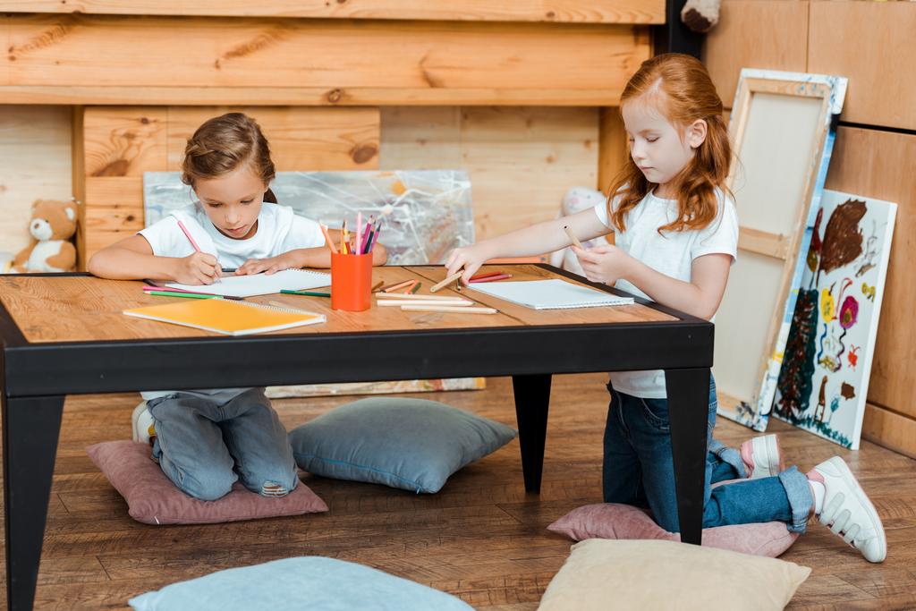 Niedliche Kinder malen auf Papieren, während sie auf Kissen sitzen  - Foto, Bild
