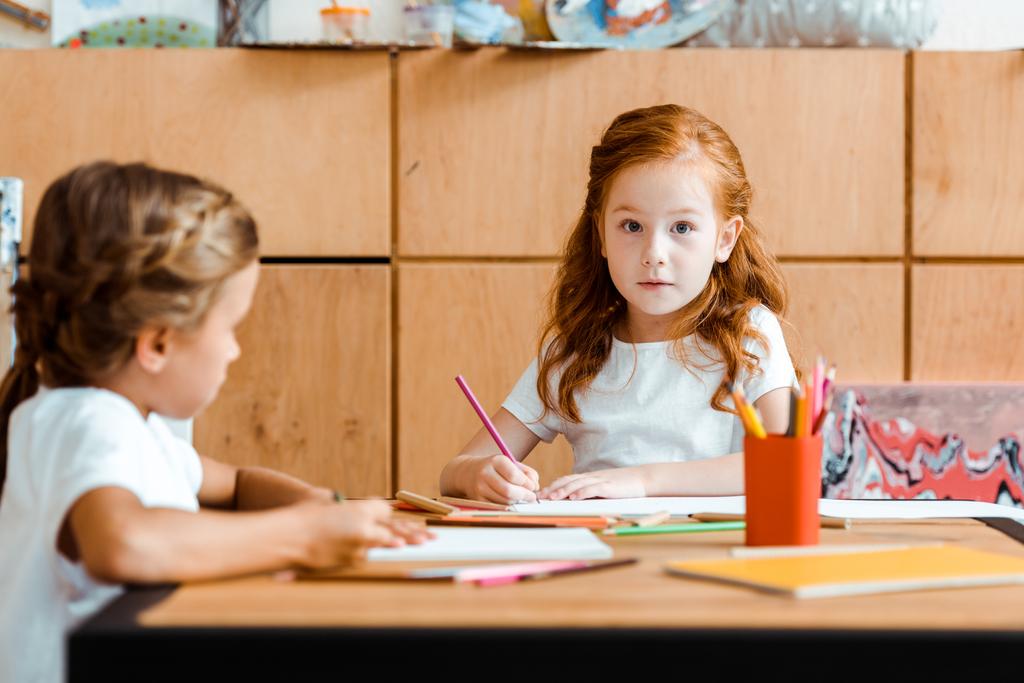 selektywna ostrość zaskoczonego rudego dziecka patrzącego na kamerę w pobliżu dziecka z kolorowym ołówkiem  - Zdjęcie, obraz