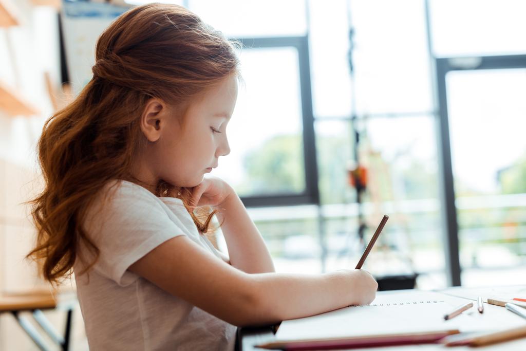 zijaanzicht van roodharige kind tekening op papier met kleurpotlood  - Foto, afbeelding
