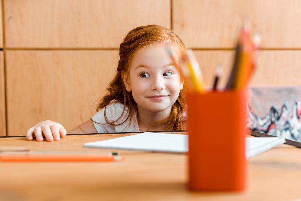 вибірковий фокус усміхненої рудої дитини біля кольорових олівців на столі
  - Фото, зображення