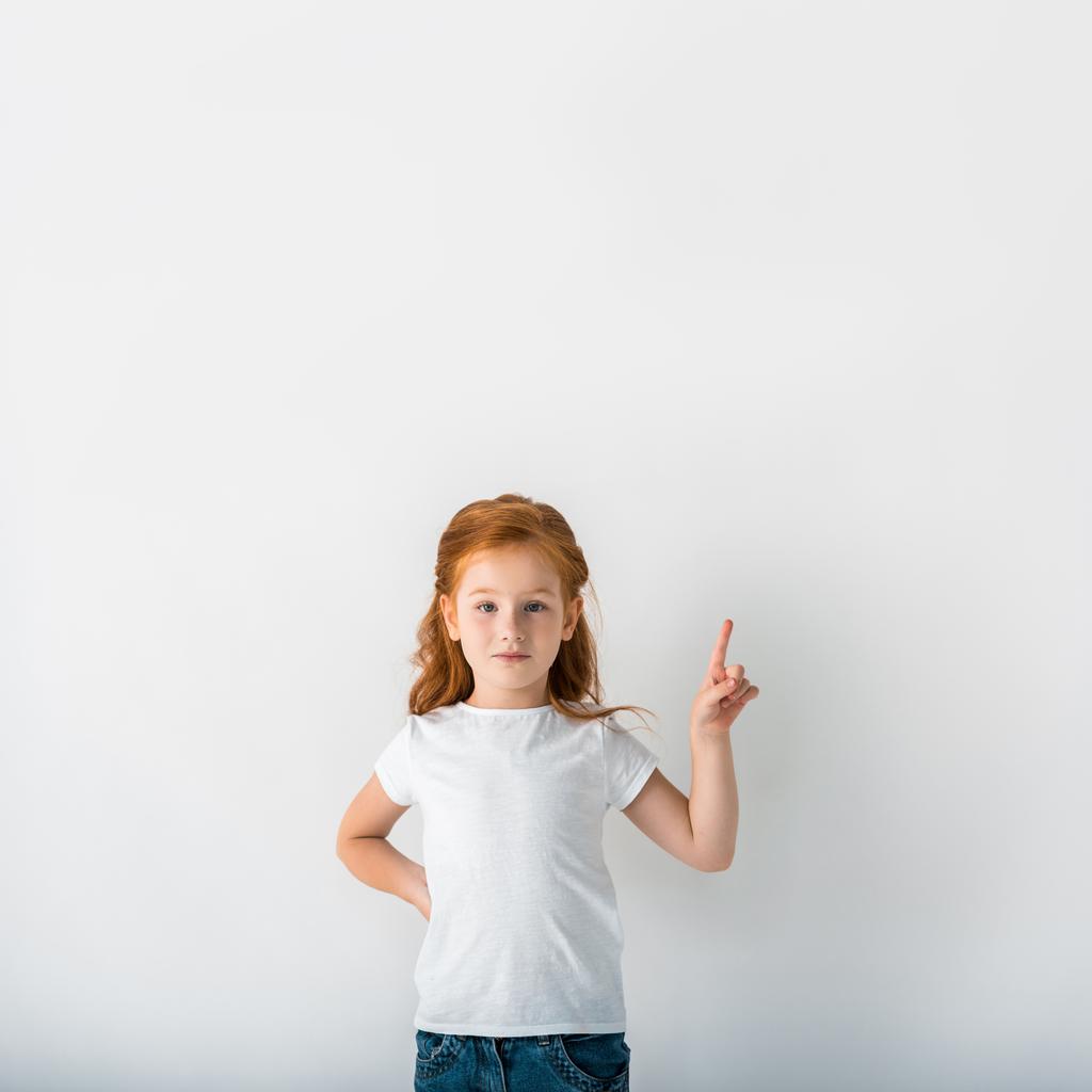 χαριτωμένο παιδί με κόκκινα μαλλιά κοιτάζοντας κάμερα και δείχνοντας με το δάχτυλο στο λευκό  - Φωτογραφία, εικόνα