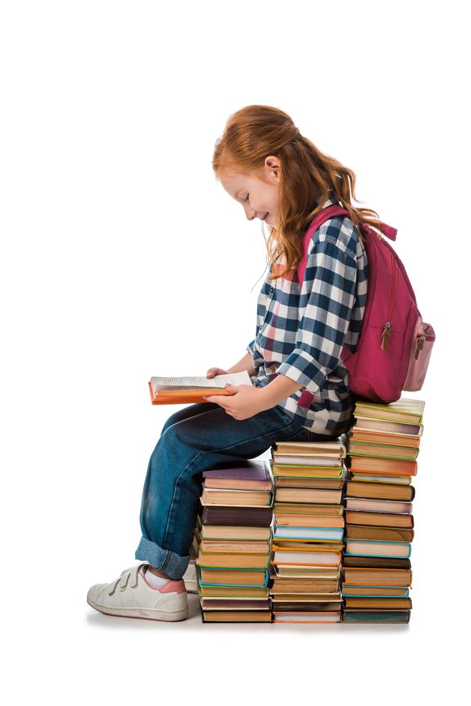 χαρούμενο κοκκινομάλλικο σχολιαρόπαιδο χαμογελά ενώ κάθεται σε βιβλία απομονωμένα στα λευκά  - Φωτογραφία, εικόνα