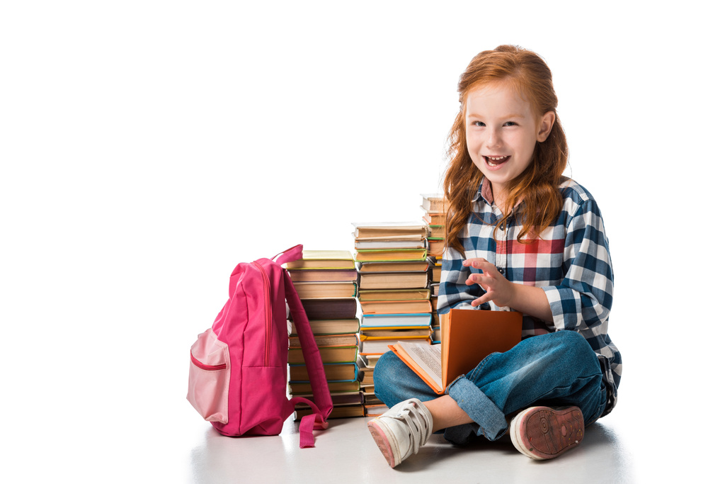vrolijk roodharige schoolkind zitten in de buurt van boeken en roze rugzak op wit  - Foto, afbeelding