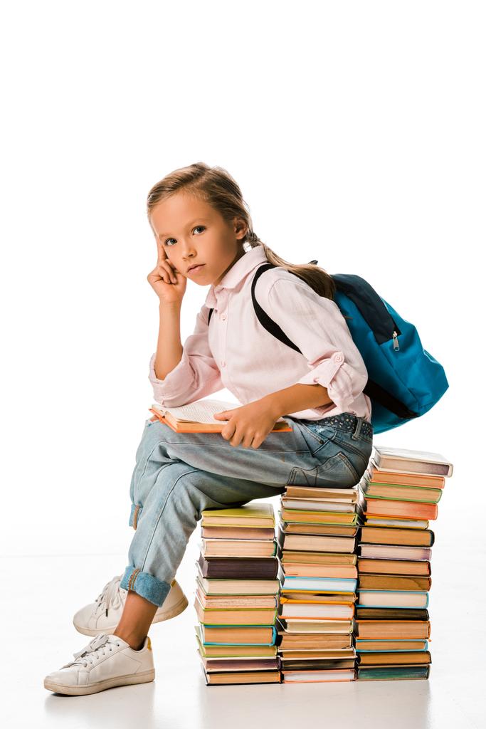 entzückendes Schulkind sitzt auf Büchern und schaut in die Kamera auf weiß  - Foto, Bild
