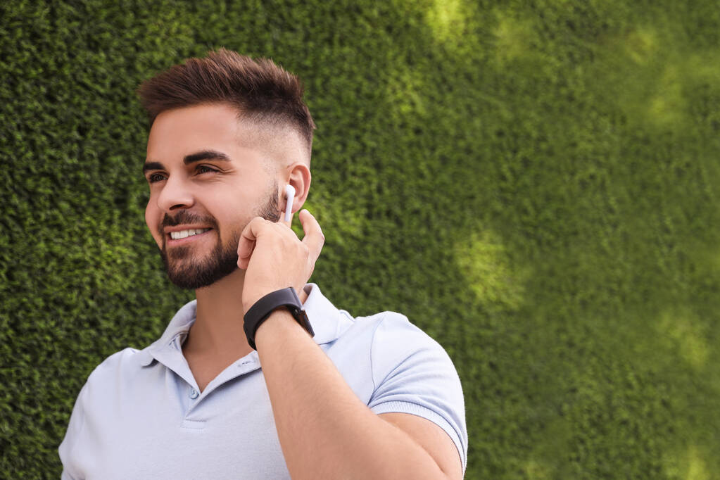Νεαρός άνδρας με ασύρματα ακουστικά ακούγοντας μουσική κοντά στο πράσινο γρασίδι τοίχο. Χώρος για κείμενο - Φωτογραφία, εικόνα