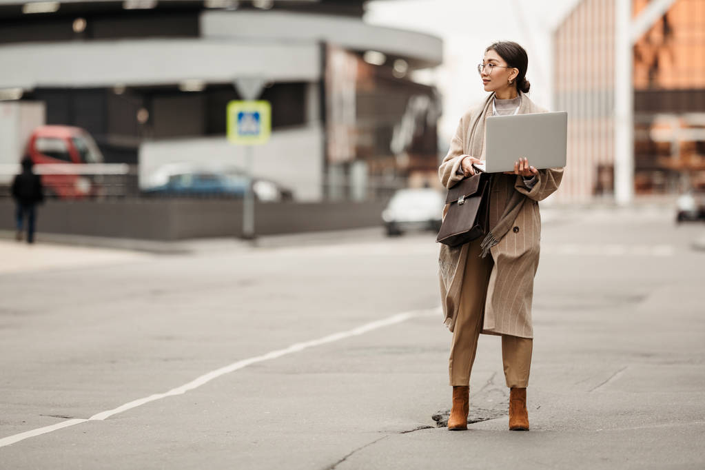 Una hermosa dama de negocios de apariencia asiática en un abrigo largo está de pie con una computadora portátil en sus manos en medio del distrito de negocios
 - Foto, imagen