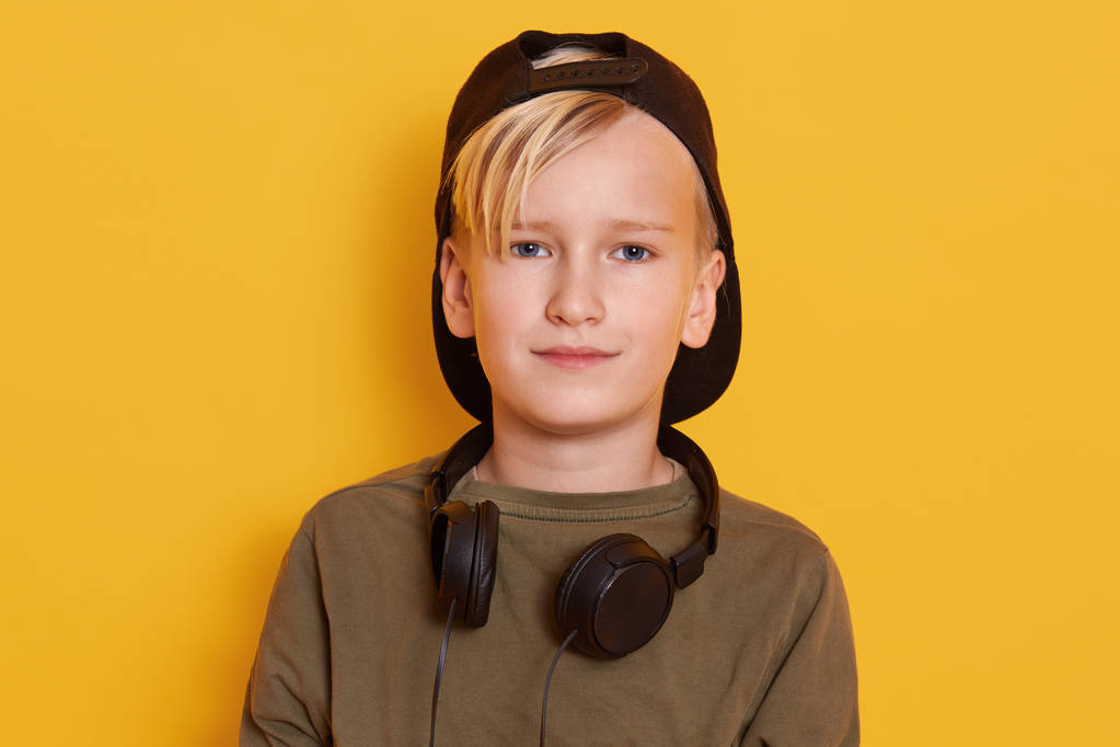 Foto Horixontal de niño rubio pequeño con auriculares alrededor del cuello, niño varón escuchando música a través de auriculares, usando camisa verde y gorra hacia atrás, posando aislado sobre fondo de estudio amarillo
. - Foto, Imagen