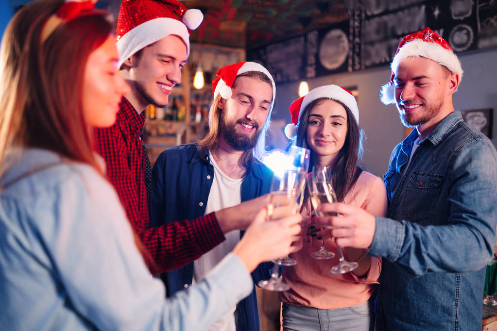 Freunde feiern gemeinsam Neujahr. Porträt von Freunden mit Drinks bei einer Cocktailparty. Die jungen Leute lachen. Gruppe schöner junger Leute mit Weihnachtsmützen. Hintergrund verschwimmen lassen. - Foto, Bild